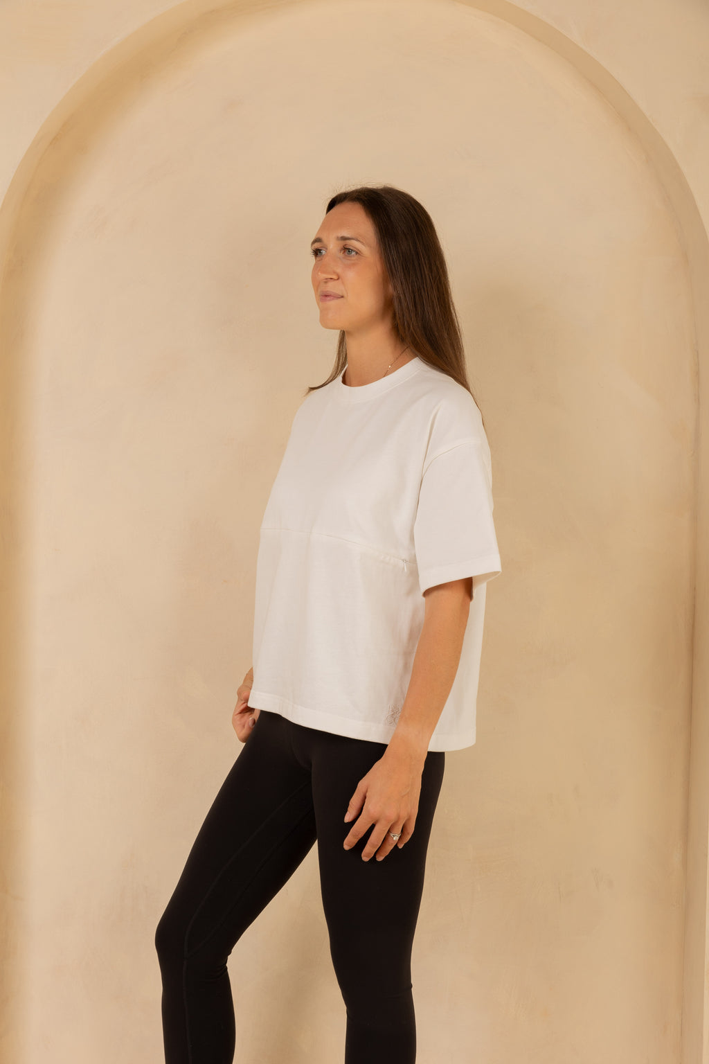 Woman wearing a white t-shirt and black pants standing against a beige wall.