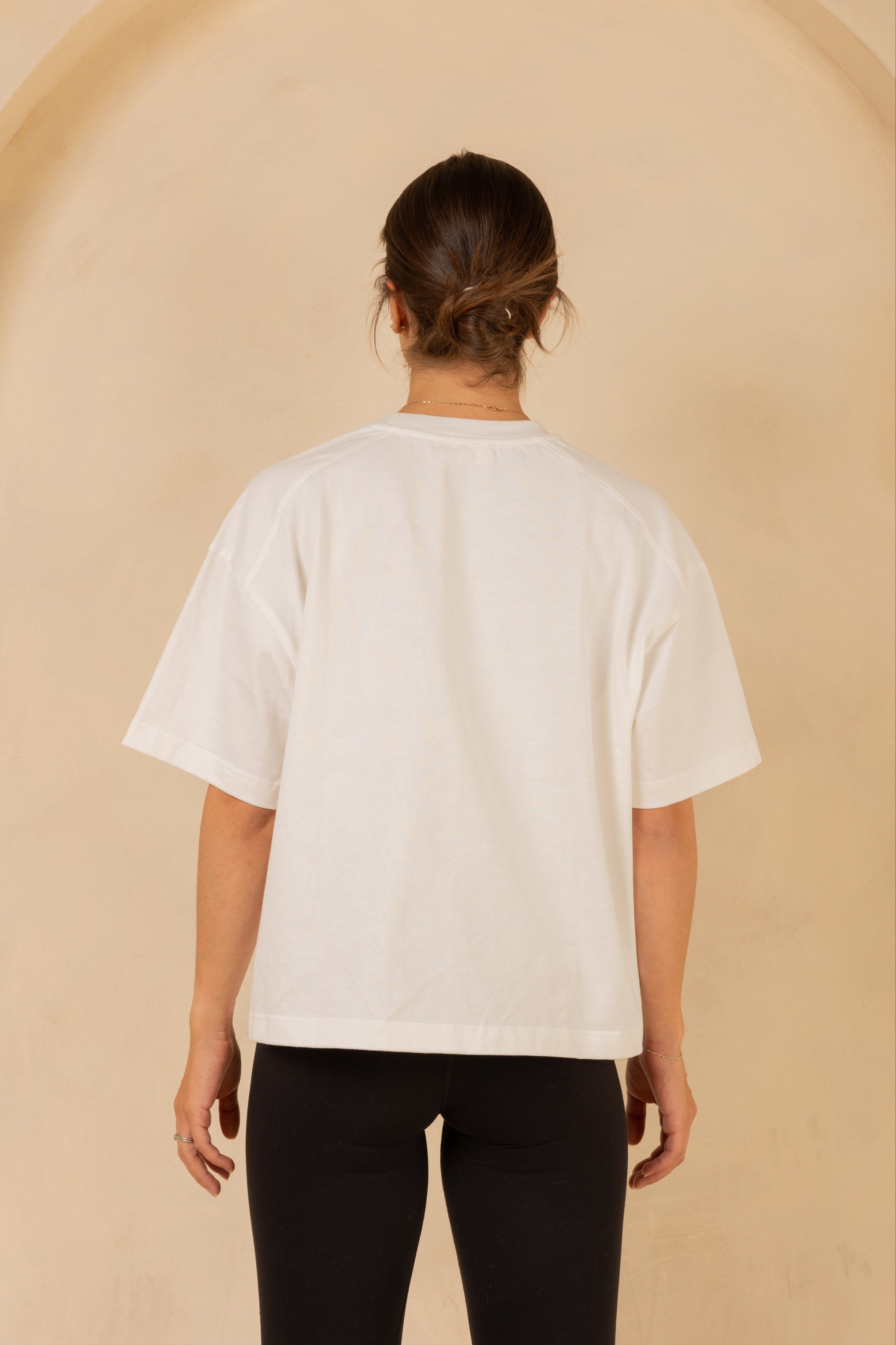 Person wearing a white t-shirt and black pants against a beige wall.